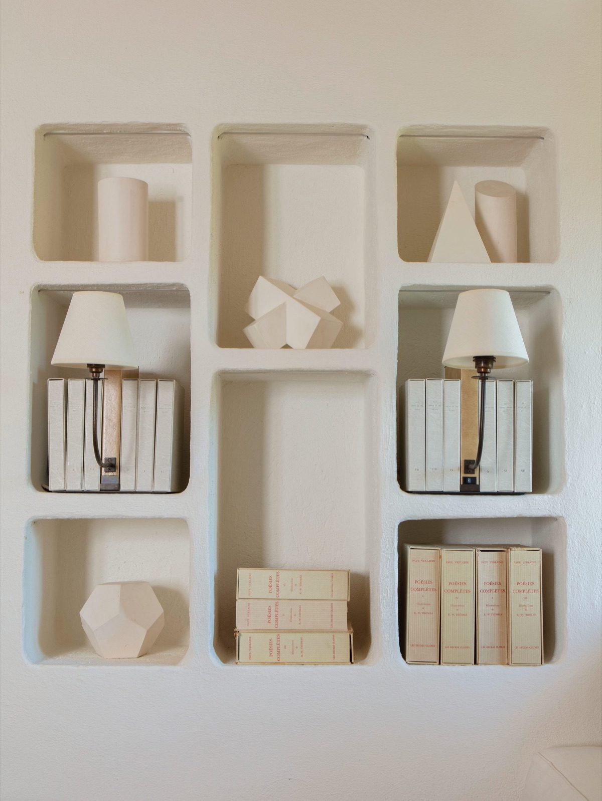 Plaster niches in a white wall, holding books, lamps and pale geometric forms
