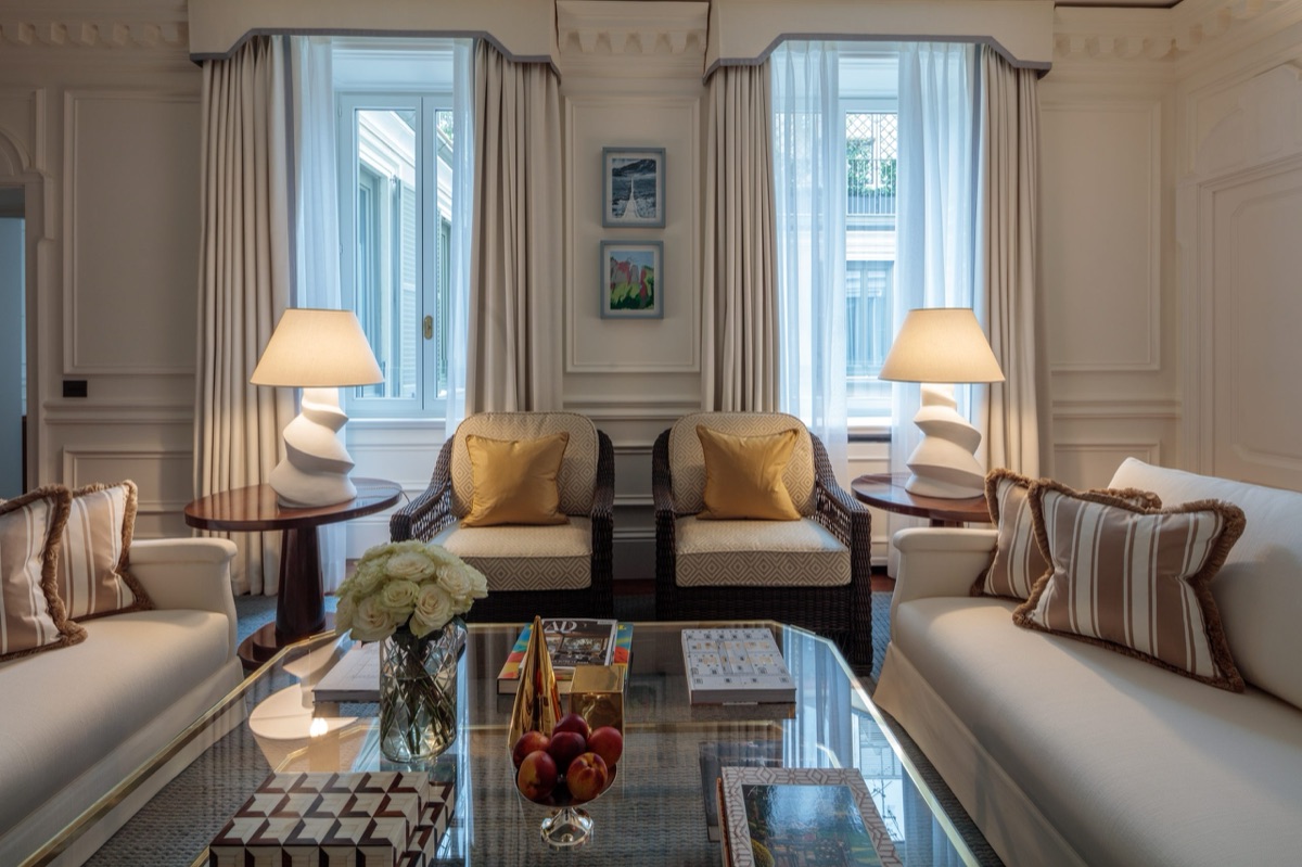 A cream sitting room with two wicker chairs, gold cushions and roses on a glass table