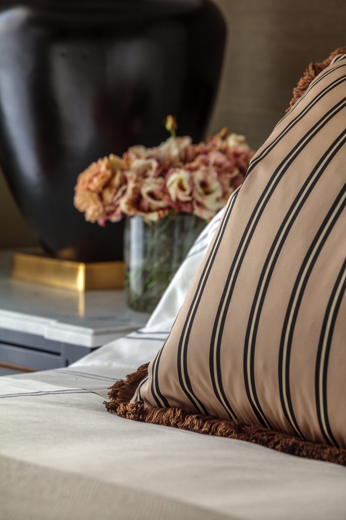 A striped bolster with a bullion fringe, lisianthus in a glass behind it
