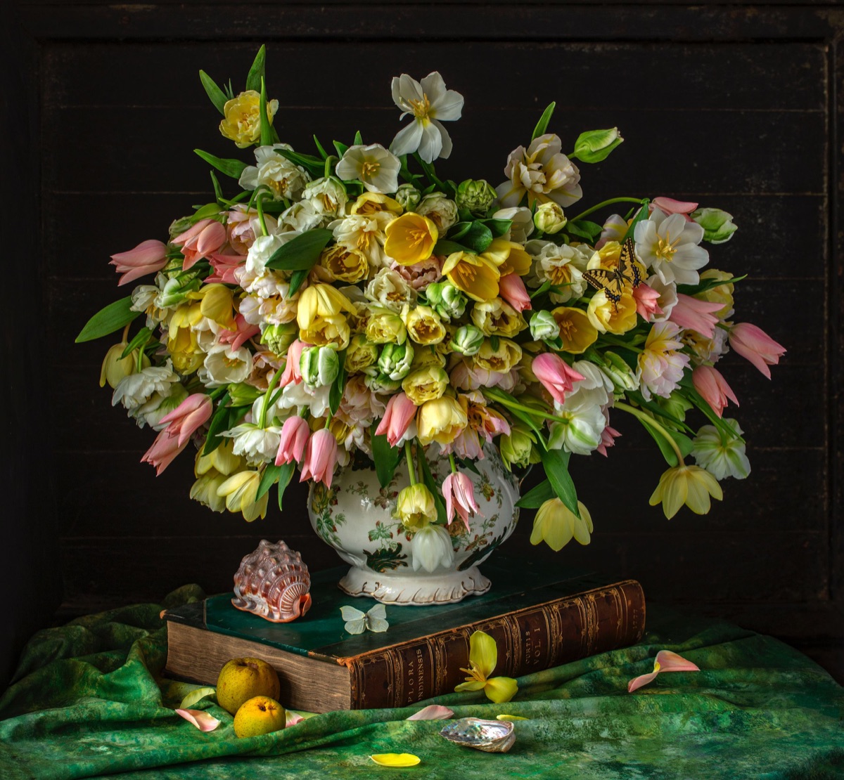 Tulips, ranunculus and narcissi in an urn on a dark table, with books and a shell