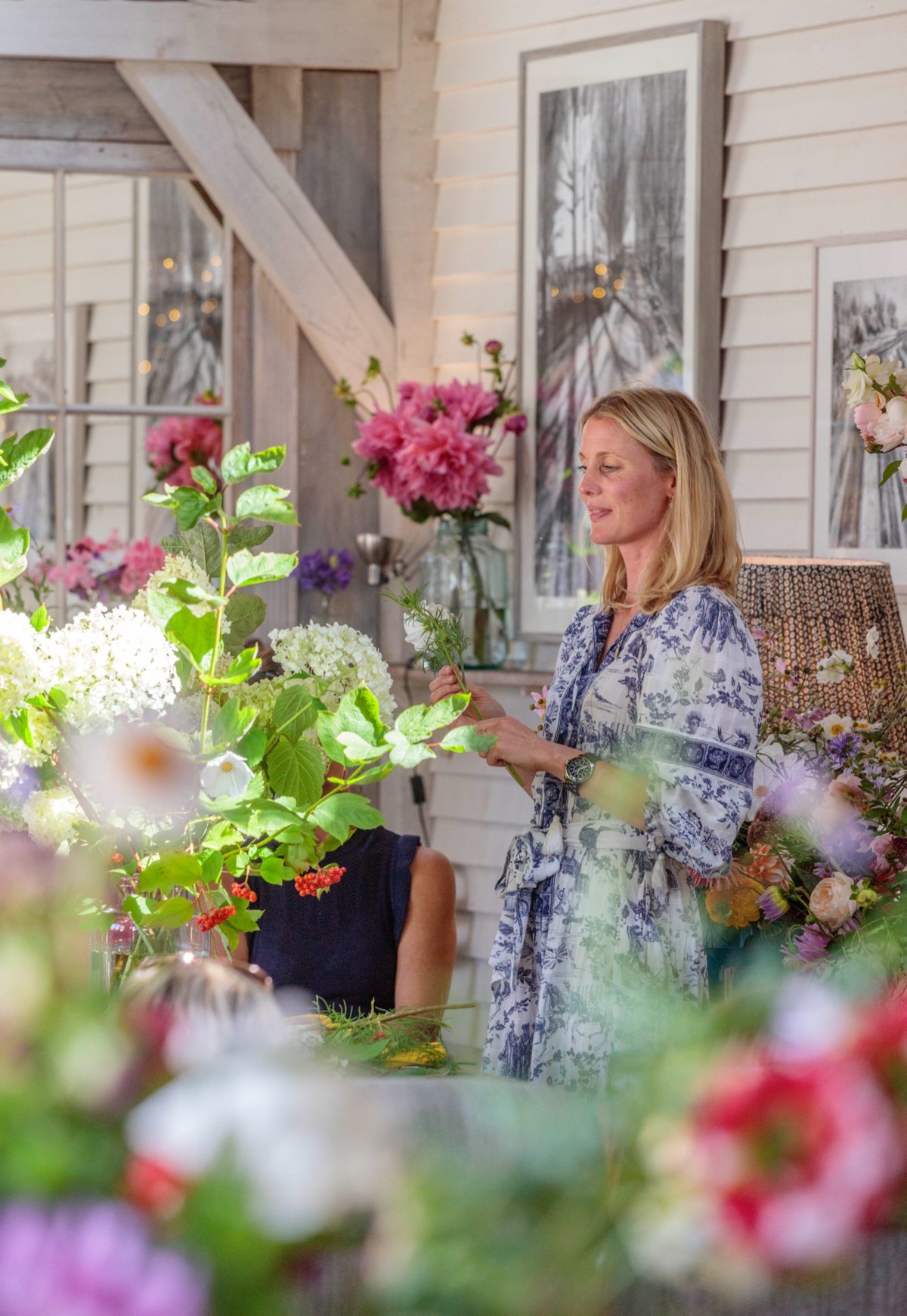 A stylist arranging cut flowers on set, dahlias and hydrangeas in the foreground, framed prints on the wall behind