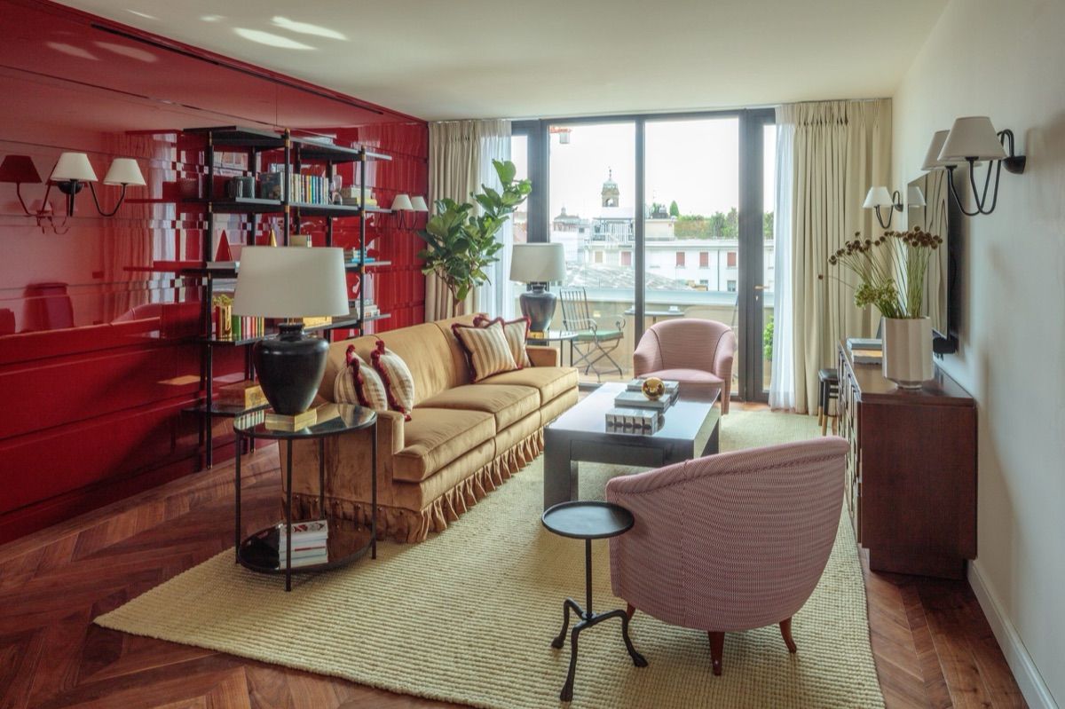 A Rocco Forte sitting room in Milan: a red lacquer wall and bookcase, a gold sofa on a jute rug, and the rooftops through the window