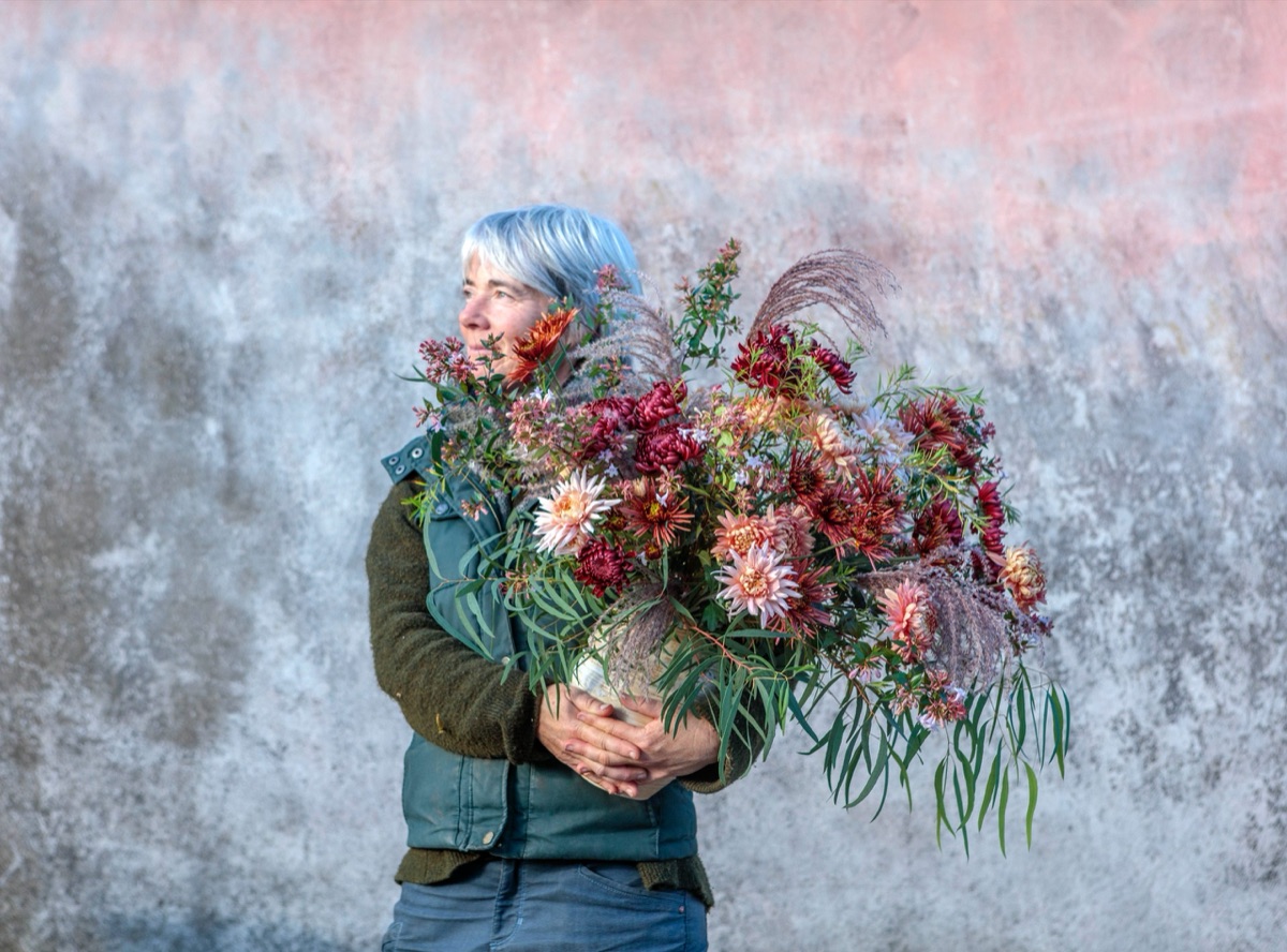 A grower with an armful of dahlias, strawflowers and grasses against a weathered wall